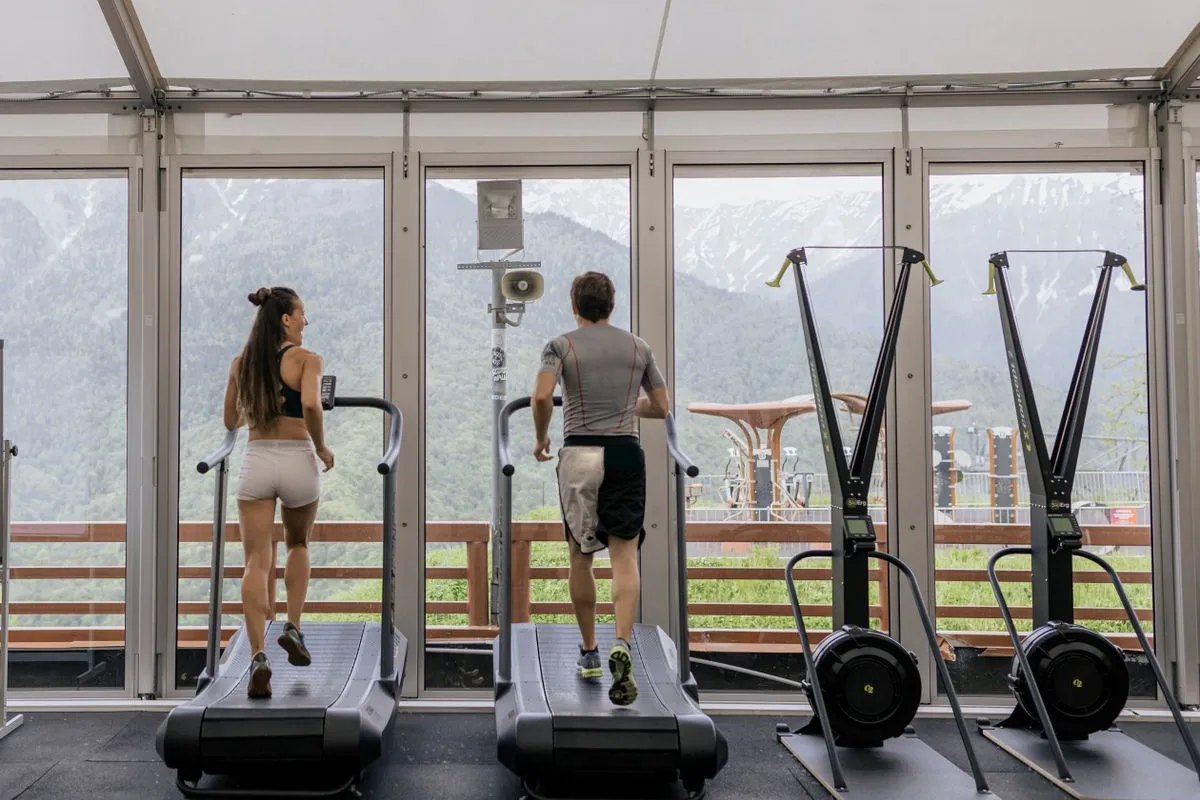 Dvoje vježbača trči na trakama u svijetloj teretani s pogledom na zelene planine