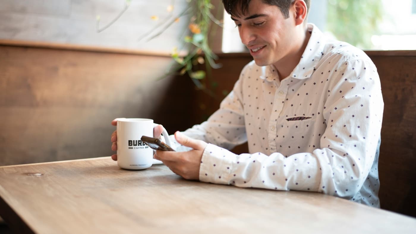 Ein Mann mit einer Tasse Kaffee an einem Tisch im Café lächelt, während er auf sein Smartphone schaut